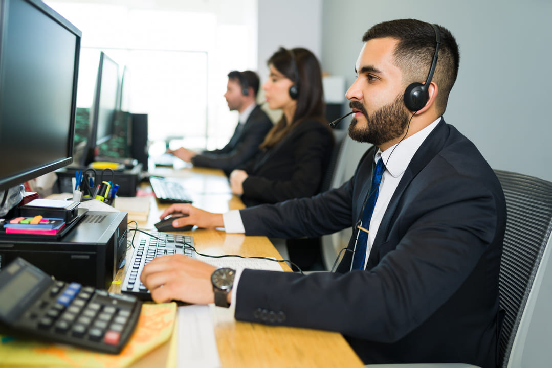 Uma equipe de atendimento está trabalhando em um ambiente corporativo. Três profissionais, vestidos formalmente com ternos e gravatas, estão sentados em uma mesa com computadores. Eles usam headsets, indicando que estão realizando ou recebendo chamadas. No primeiro plano, um homem de barba, vestido com um terno escuro e gravata azul, está focado em seu monitor. A cena ocorre em um escritório iluminado, com papéis, calculadora e materiais de trabalho visíveis na mesa.