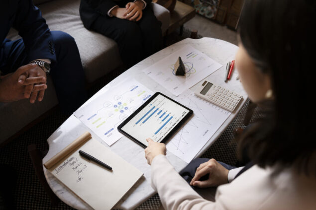 Três pessoas analisam gráficos e relatórios financeiros em uma mesa com tablet, papéis, calculadora e anotações.