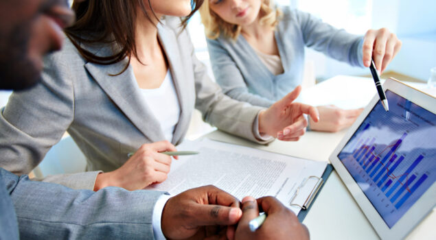 Um grupo de três profissionais de negócios está discutindo dados apresentados em um tablet. Uma mulher à direita aponta para um gráfico de barras e linhas na tela, enquanto outra mulher ao centro escuta atentamente e segura uma caneta. Um homem no primeiro plano está tomando notas em uma prancheta com documentos. A atmosfera é iluminada por luz natural, sugerindo um ambiente de escritório moderno e colaborativo.
