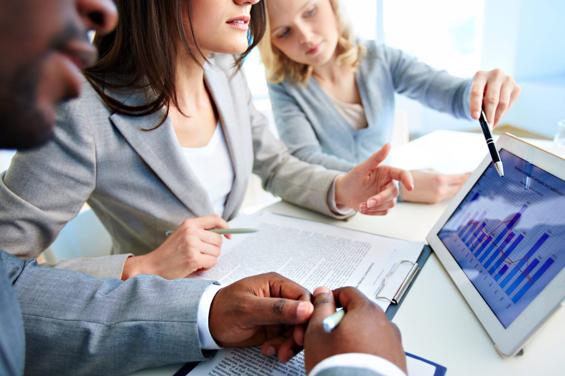 Um grupo de três profissionais de negócios está discutindo dados apresentados em um tablet. Uma mulher à direita aponta para um gráfico de barras e linhas na tela, enquanto outra mulher ao centro escuta atentamente e segura uma caneta. Um homem no primeiro plano está tomando notas em uma prancheta com documentos. A atmosfera é iluminada por luz natural, sugerindo um ambiente de escritório moderno e colaborativo.