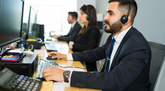 Uma equipe de atendimento está trabalhando em um ambiente corporativo. Três profissionais, vestidos formalmente com ternos e gravatas, estão sentados em uma mesa com computadores. Eles usam headsets, indicando que estão realizando ou recebendo chamadas. No primeiro plano, um homem de barba, vestido com um terno escuro e gravata azul, está focado em seu monitor. A cena ocorre em um escritório iluminado, com papéis, calculadora e materiais de trabalho visíveis na mesa.