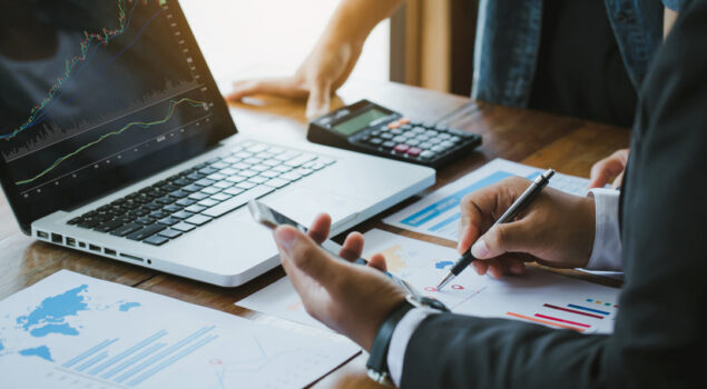 Duas pessoas analisando gráficos financeiros impressos sobre uma mesa de madeira, com um notebook exibindo gráficos de ações, uma calculadora e documentos ao redor.
