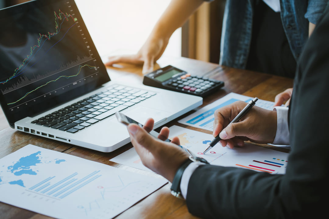 Duas pessoas analisando gráficos financeiros impressos sobre uma mesa de madeira, com um notebook exibindo gráficos de ações, uma calculadora e documentos ao redor.