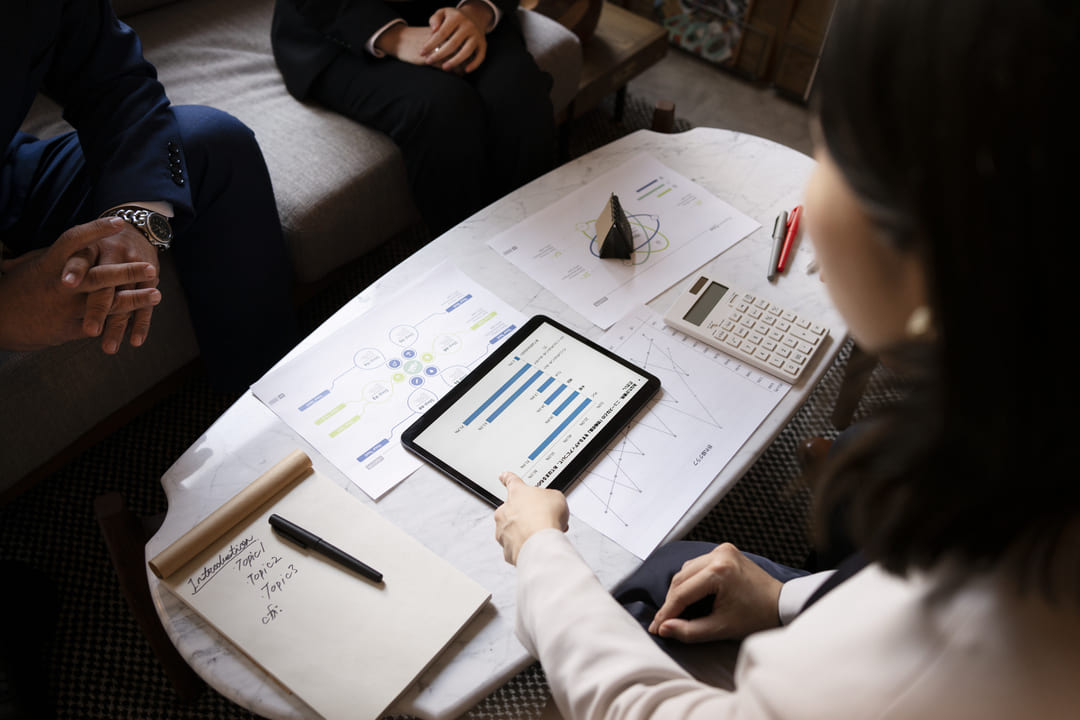 Três pessoas analisam gráficos e relatórios financeiros em uma mesa com tablet, papéis, calculadora e anotações.