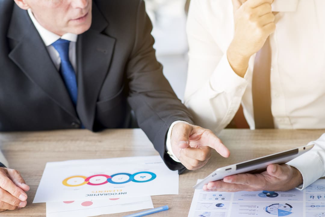 Dois homens de negócios em reunião analisam relatórios impressos e dados em um tablet, discutindo informações relacionadas à controladoria estratégica.