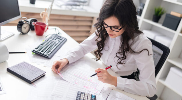 Mulher trabalhando em escritório, revisando documentos e planilhas financeiras sobre a mesa, representando organização contabilidade.