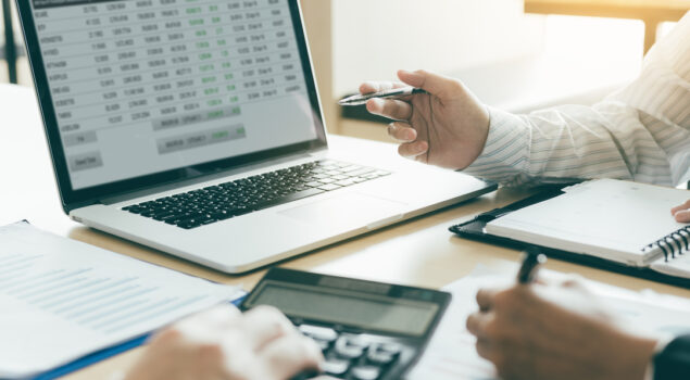 Duas pessoas analisando planilhas financeiras no computador, com calculadora e anotações em papel sobre a mesa.