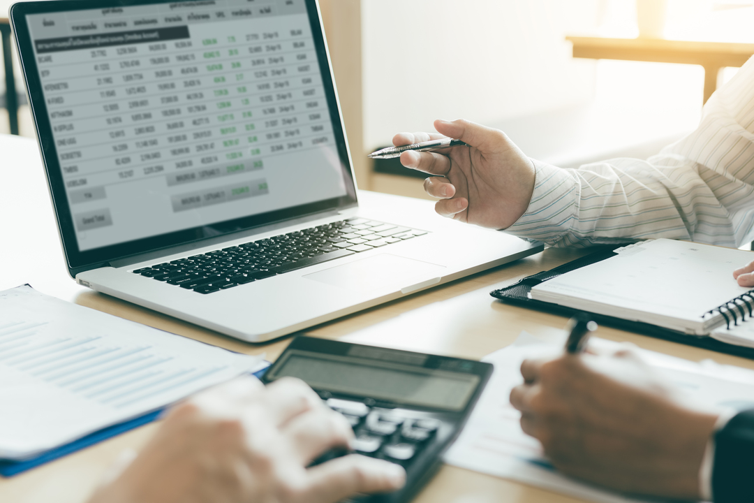 Duas pessoas analisando planilhas financeiras no computador, com calculadora e anotações em papel sobre a mesa.