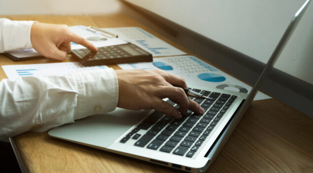 Pessoa utilizando um notebook e uma calculadora sobre uma mesa com relatórios financeiros, representando a automação e o controle da folha de pagamento terceirizada.