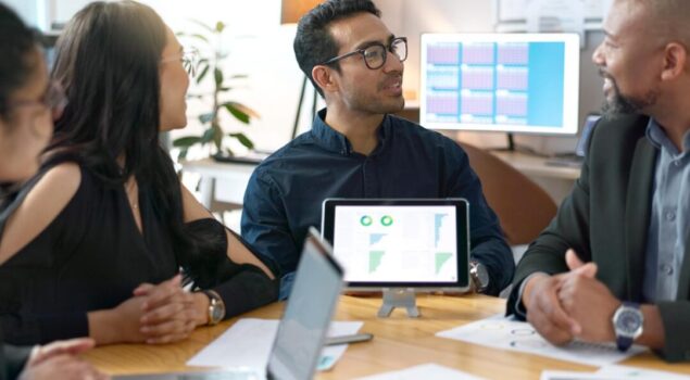 Equipe em reunião analisando gráficos e indicadores exibidos em um tablet e computadores.