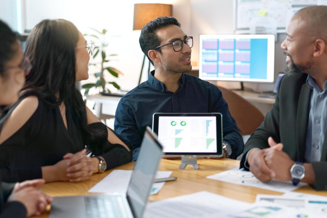 Equipe em reunião analisando gráficos e indicadores exibidos em um tablet e computadores.