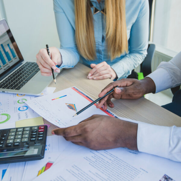 Profissionais analisam relatórios financeiros e gráficos em uma mesa de trabalho, com notebook e calculadora.