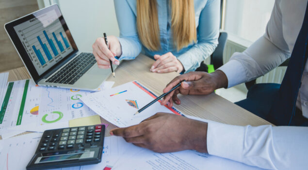 Profissionais analisam relatórios financeiros e gráficos em uma mesa de trabalho, com notebook e calculadora.